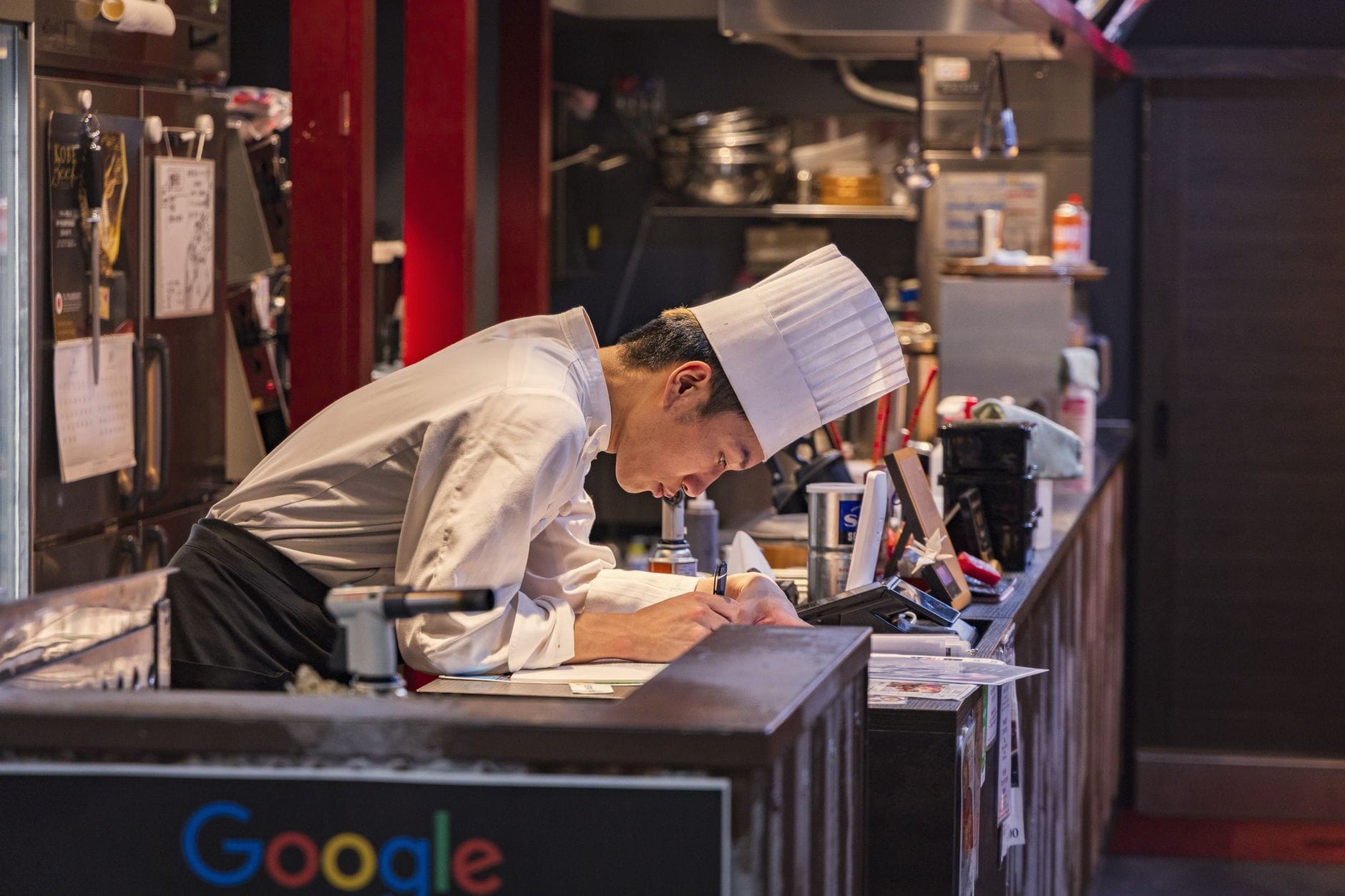 Chef in a kitchen, focused on writing with culinary equipment around.